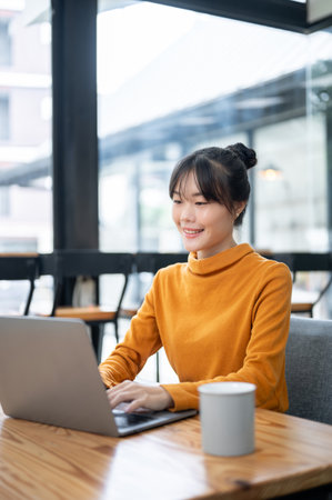 A charming, positive Asian woman is working remotely from a coffee shop, working on her laptop computer at a table. freelance, digital nomad, businesswoman, college studentの写真素材