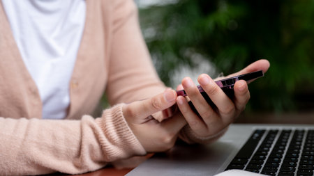 A close-up image of a woman in a cardigan is using her smartphone while working remotely at a table in a garden, a laptop on the table. people and technology conceptsの写真素材