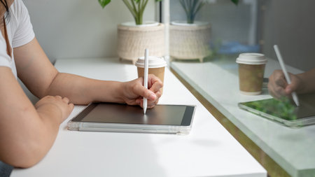 A close-up side view image of a woman sitting at a table against the window, holding a stylus pen,...
