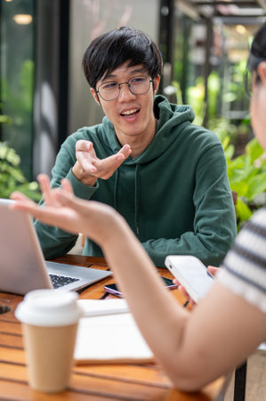 A young Asian man in a green hoodie is talking, discussing work with his friend while sitting at a table outdoors. brainstorming, consulting, teamworkの写真素材