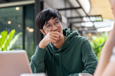A young happy Asian male college student in a green hoodie is enjoying talking with his friend while doing homework together at a cafe.の写真素材