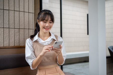 A happy, beautiful Asian woman is enjoying chatting with her friends on her smartphone while relaxing sitting indoors. people and wireless technology conceptsの写真素材