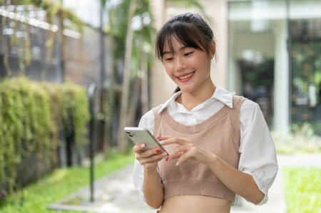A beautiful, confident Asian woman is using her smartphone while standing outdoors. chatting, reading text messages, scrolling on the phone, shopping onlineの写真素材