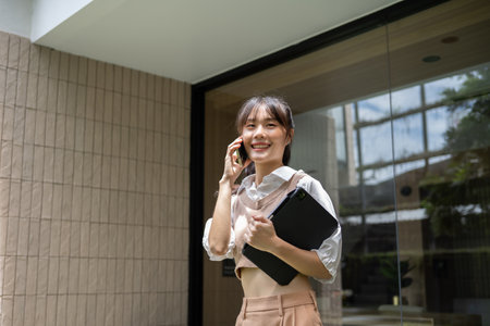 A beautiful, positive Asian woman is talking on the phone while walking out the building, holding a digital tablet, standing in front of the entrance door.の写真素材