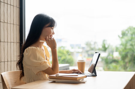 A side view image of a beautiful Asian woman working on her digital tablet at a table in a room, looking at the tablet screen.の写真素材