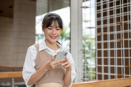 A beautiful, happy young Asian woman in casual clothes is texting someone or scrolling on her smartphone while standing in the building. people and wireless technology conceptsの写真素材