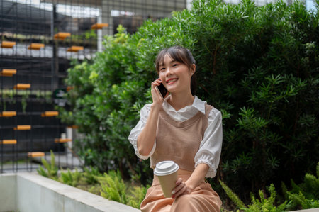 An attractive, happy Asian woman is having coffee and talking on the phone while resting outdoors during her lunch break.の写真素材