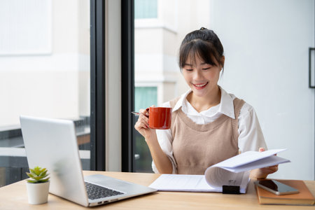 A beautiful Asian businesswoman having a morning coffee while reviewing business report at her desk, working in her modern office.の写真素材