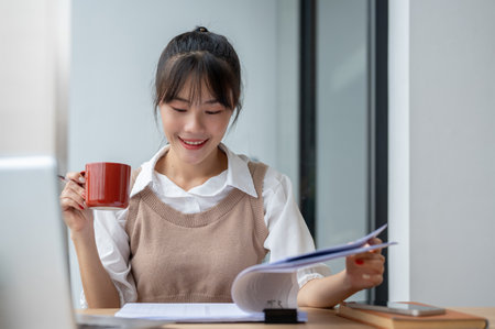 A beautiful, positive Asian businesswoman is having her morning coffee while reviewing business paperwork and working at her desk in the office.の写真素材
