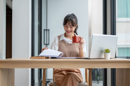 A beautiful, positive Asian businesswoman is having her morning coffee while reviewing business paperwork and working at her desk in the office.の写真素材