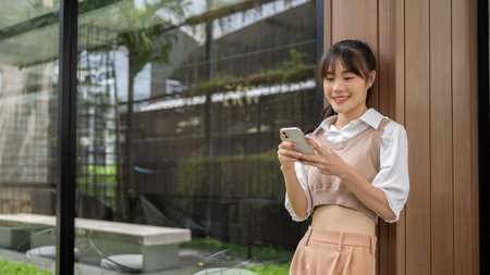 A positive, charming Asian woman is leaning against the wall outdoors, using her smartphone, chatting, or scrolling on the phone. people, wireless technology, lifestyleの写真素材