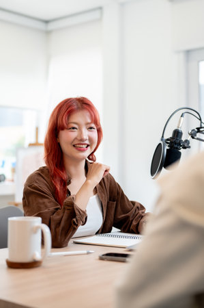 A confident and friendly Asian female podcaster is enjoying a conversation as she interviews a guest in her studio, recording a podcast with a special guest.の写真素材