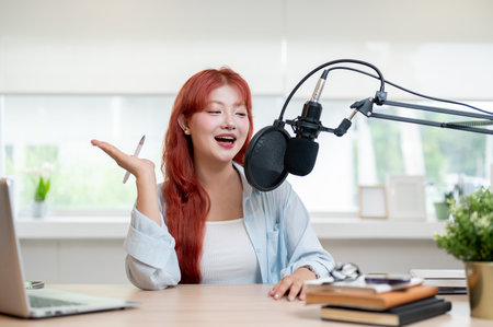 A charming and cheerful Asian female podcaster with red curly hair is speaking into a microphone, enjoying the process of recording her podcast in the studio.の写真素材