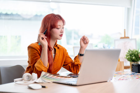 A focused and creative Asian female graphic designer with red hair is working at her studio table, which is equipped with a laptop and a set of color swatches.の写真素材