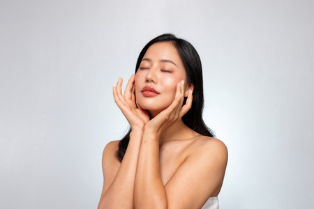 A young Asian beauty with a gorgeous face and glowing tanned skin poses against a grey background, gently touching her face with her eyes closed, lost in a dreamy expression. facial treatment cosmeticの写真素材
