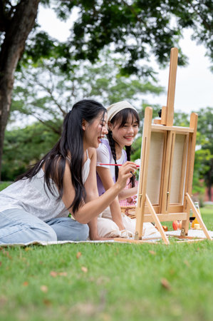 Two positive and cute Asian best friends are enjoying painting on a canvas easel together while picnicking in a green park on a bright summer day. leisure and friendship conceptsの写真素材