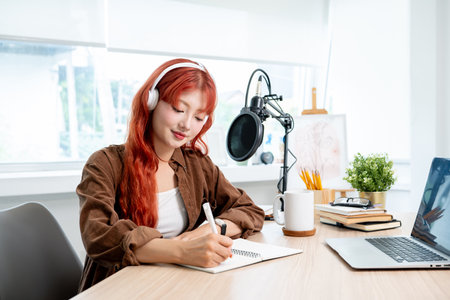 A charming and positive Asian female radio host or journalist with red curly hair is working in her studio, focusing on adjusting the script for her show.の写真素材