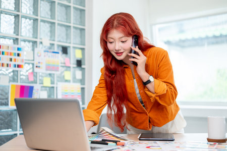 A confident Asian female graphic designer with red curly hair is talking on the phone while leaning against the table and working on her laptop, working in her studio.の写真素材