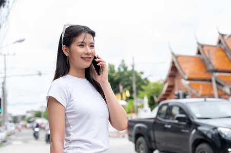 A beautiful, positive young Asian woman is talking on the phone while standing by the street in the old town city of Chiang Mai, Thailand. people, lifestyles, wireless technologyの写真素材