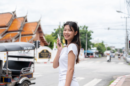 A beautiful, positive young Asian woman is talking on the phone while standing by the street in the old town city of Chiang Mai, Thailand, smiling at the camera. people, wireless technologyの写真素材