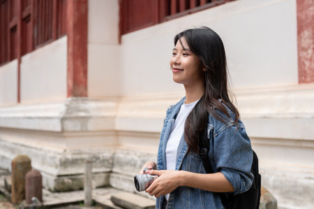 A beautiful, smiling Asian female tourist in a denim jacket and backpack, holding a camera, is visiting a stunning Thai temple in Chiang Mai. Asian traveler, vacation, solo travelingの写真素材
