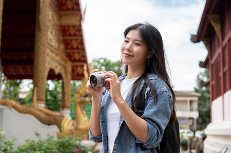 A beautiful, smiling Asian female tourist in a denim jacket and backpack, holding a camera, is visiting a stunning Thai temple in Chiang Mai. Asian traveler, vacation, solo travelingの写真素材