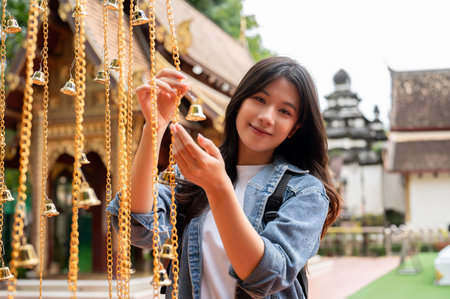 A beautiful, positive Asian female tourist is hanging a bell for luck in a temple during her visit...