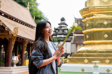 A positive, charming young Asian female tourist in a denim jacket is putting her hands together in a prayer position with a flower, making a wish while visiting a temple in Thailand.の写真素材
