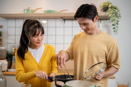 A pretty and positive young Asian woman is helping her boyfriend in the kitchen as they cook together, sharing a happy moment at home. home cooking, domestic life, couplesの写真素材