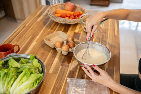 A close-up image of a woman is whisking pancake mix batter in a bowl at the kitchen table while...
