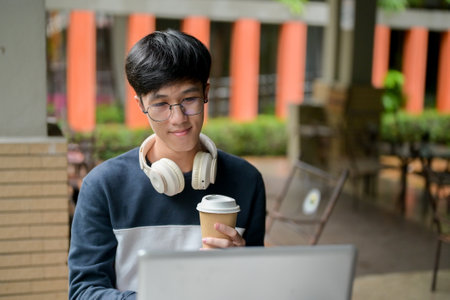 A young, positive Asian male college student wearing glasses is working on his laptop and having...