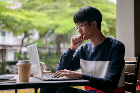 A thoughtful young Asian man is working remotely from an outdoor table, working on his laptop computer, reading online news or emails. people and technology conceptsの写真素材