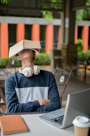 A tired, sleepy young Asian male college student rests with a book covering his face while working...