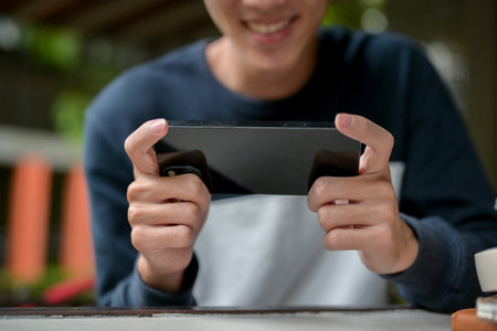 A cropped image of a joyful young Asian man happily playing a mobile game on his smartphone while sitting at a table. people and wireless technology conceptsの写真素材