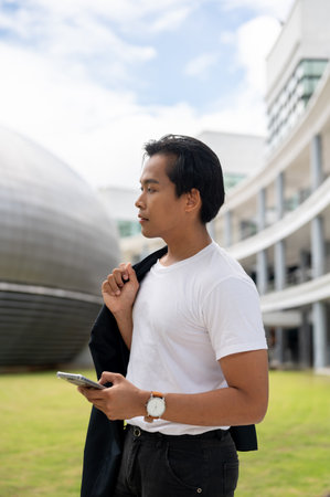 A portrait of a good-looking Asian man holding his suit and standing outdoors with a smartphone in his hand, looking away, with modern buildings in the background.の写真素材
