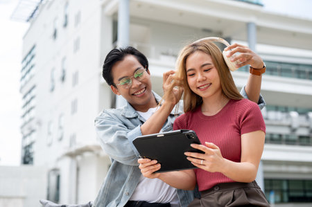 A caring, lovely young Asian man is placing headphones on his girlfriend while she looks at a tablet, as they sit outdoors on campus with modern buildings in the background.の写真素材