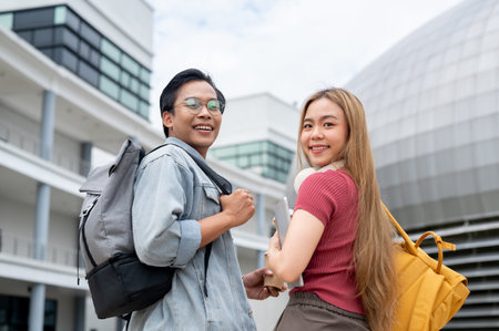 Two cheerful young Asian college students, a male and a female, in casual clothes and backpacks, are smiling at the camera while standing outdoors with modern college buildings in the background.の写真素材