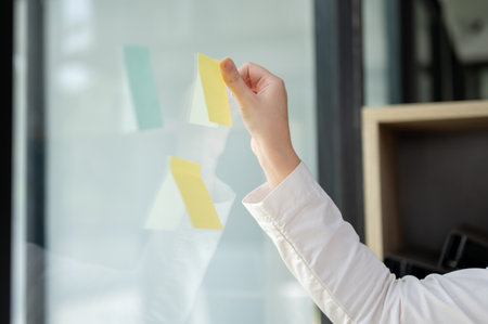 A close-up image of a businesswoman attaching a sticky note to a glass wall in the office. brainstorming idea and co-working conceptsの写真素材