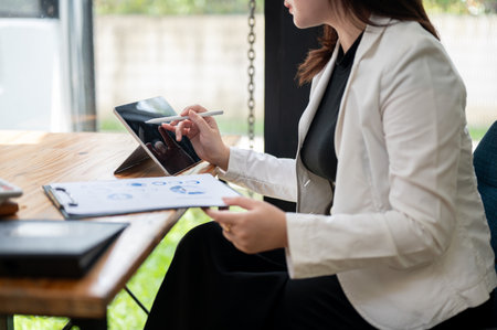 A cropped side view image of a businesswoman in a white business suit working on her digital tablet and paperwork at her desk in the office. businesspeople and wireless technology conceptsの写真素材