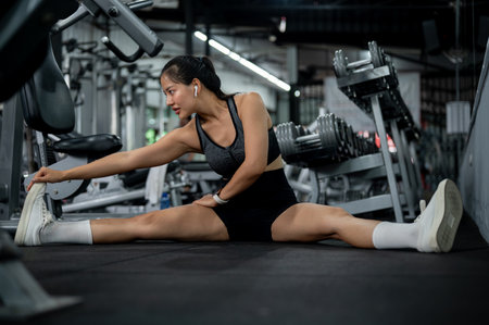 An attractive, sporty Asian woman in athletic wear is sitting on the gym floor, stretching her legs,...