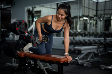 An attractive, strong, and fit Asian woman in athletic wear is performing dumbbell tricep kickbacks...