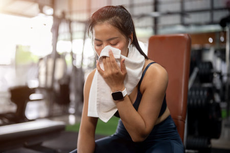 A tired, sporty Asian woman in athletic wear sits on a bench press to rest after her workout in the gym, wiping sweat from her face with a towel. workout, fitness training, exerciseの写真素材