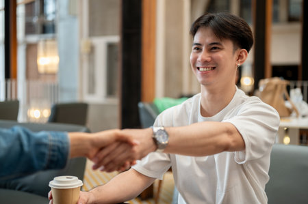 A happy, handsome young Asian man giving a handshake to his friend during a meeting at a coffee shop. friendship, companion, teamwork, support, togethernessの写真素材