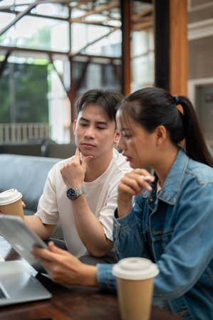 Two young Asian friends, a male and a female, are working remotely together from a coffee shop, looking at a tablet screen, discussing work and planning a project.の写真素材