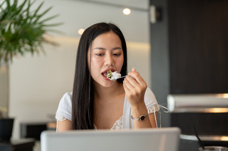 An attractive Asian woman is eating cake while focusing working on her laptop in a coffee shop, working remotely. city life, modern lifestyles, wireless technologyの写真素材