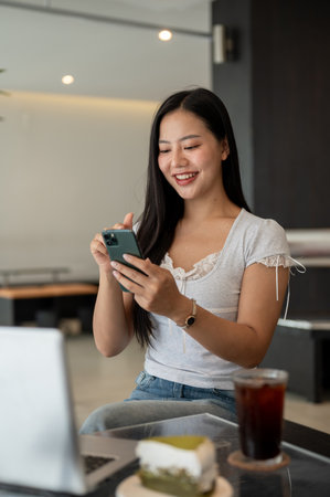 A beautiful, positive Asian woman using her smartphone while sitting in a modern coffee shop. reading text, replying chat, sending messages, scrolling on the phone, browsing webの写真素材