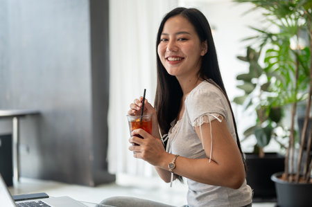 A side view of a beautiful Asian woman with an iced americano in her hand is smiling at the camera, sitting in a modern coffee shop, working remotely.の写真素材