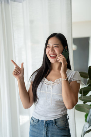 An attractive, cheerful Asian woman is enjoying talking on the phone with someone while standing by the window indoors. people and wireless technology conceptsの写真素材