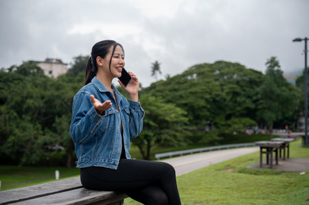A positive Asian woman in a denim jacket is talking on the phone with someone on a bench in a park. people, lifestyle, wireless technologyの写真素材