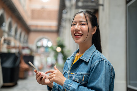 A beautiful, positive Asian woman in a denim jacket stands outdoors with a building in the background, holding a smartphone and smiling while looking away. people, lifestyle, wireless technologyの写真素材